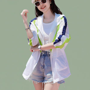 Woman wearing a white jacket with green and blue accents, sunglasses, and denim shorts by the water.