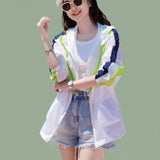 Woman wearing a white jacket with green and blue accents, sunglasses, and denim shorts by the water.